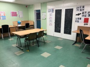 A classroom with green tile flooring, light green walls, and wooden tables and black chairs. Chalkboard drawings are on the doors, and an educational poster is on the wall. A globe sits on a side table, conveying an educational tone.