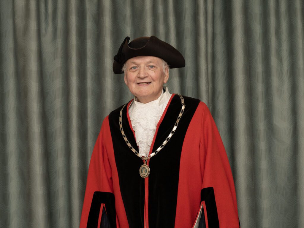 Councillor Ray Perry - Mayor and Chair of Yate An older person wearing a tricorn hat and red ceremonial robe with a chain of office stands smiling against a grey curtain backdrop.