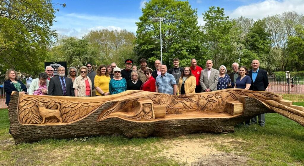A diverse group of people poses cheerfully behind a large, intricately carved wooden bench in a park. The bright day and lush trees create a lively atmosphere.