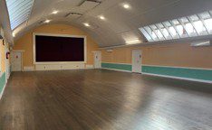 A spacious, empty hall with wooden flooring, a curved white ceiling, skylights, and a large maroon stage curtain on the far wall. The room feels bright and airy.