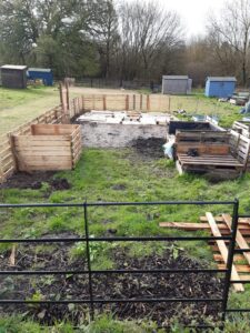A garden with wooden pallet compost bins and black metal fencing in a grassy area. Several sheds are visible in the background on a cloudy day.