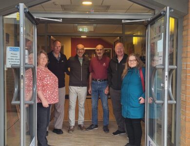A group of six people stands smiling at the entrance of a brick building with glass doors. Signs indicate a clubhouse and reception area. The mood is friendly.