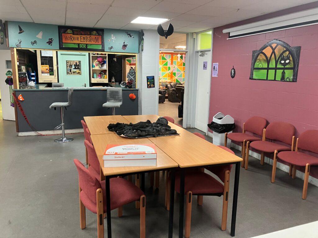 Community room decorated for Halloween with a "Horror Emporium" sign and spooky decals. Chairs line the walls; a table holds a black cloth and pizza box.
