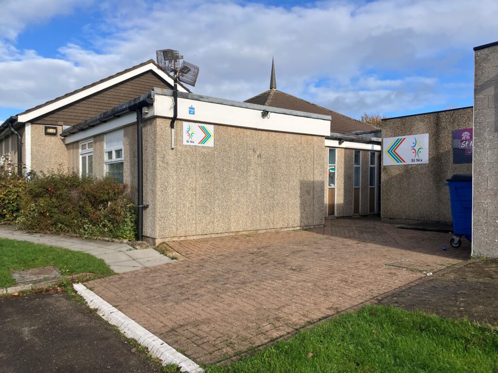 Community building with beige textured walls, colorful signs, and a blue recycling bin. The area is sunny with a partly cloudy sky and green shrubs.