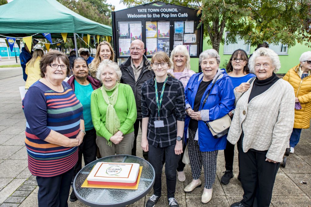 A group of smiling people stands outside under a green tent, with a cake on a table in front. A sign reads "Welcome to the Pop Inn Café."
