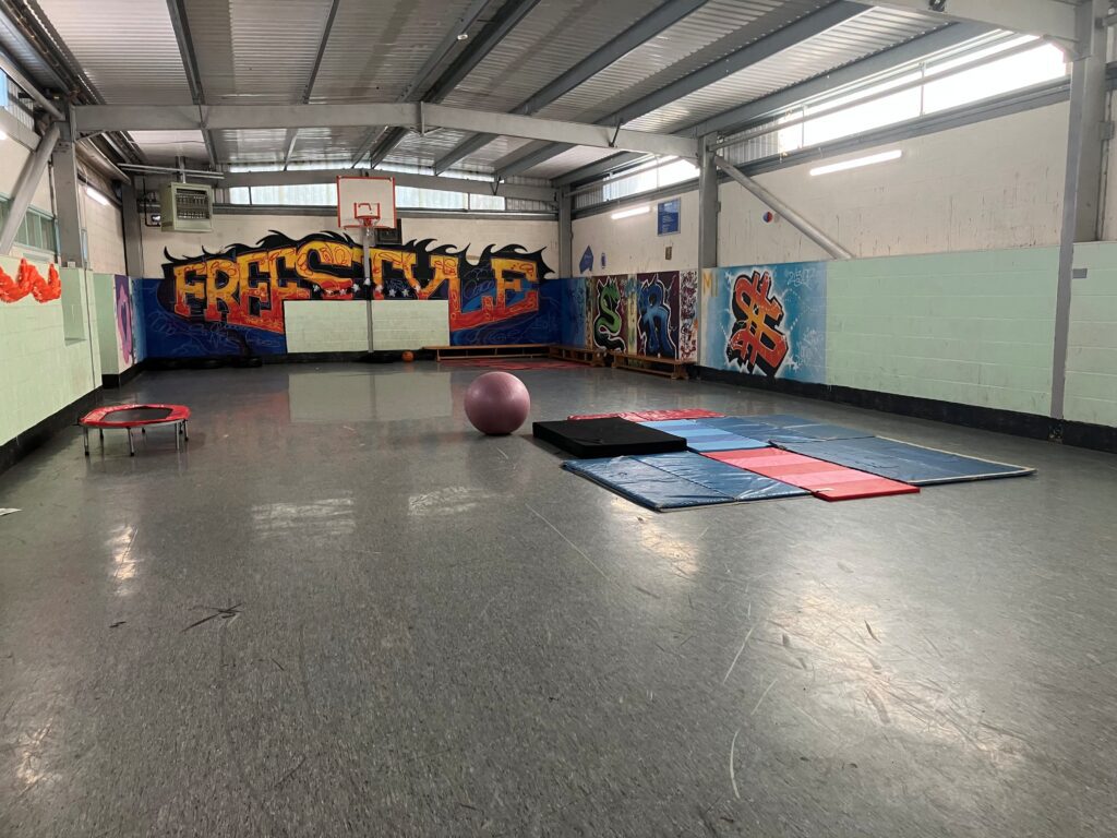 Spacious gym with graffiti walls, featuring the word "FREESTYLE." A basketball hoop, trampoline, exercise ball, and mats are visible on the floor.